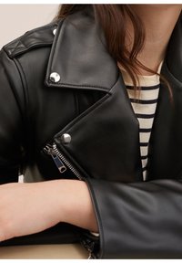 Veste de moto en cuir synthétique noir avec des détails en métal argenté, poches zippées et col structuré. Porte une chemise rayée en dessous.