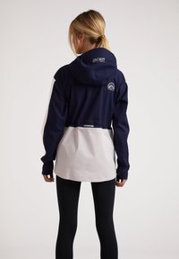 Chaqueta bloqueada en colores azul marino y blanco, con capucha, puños elásticos y dobladillo ajustable. Presenta un parche con el logo y está hecha de un tejido transpirable.