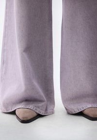 Φαρδιά παντελόνια σε ανοιχτό μωβ από τζιν ύφασμα, συνδυασμένα με μπεζ γόβες με μυτερή μύτη. Το ύφασμα παρουσιάζει απαλή υφή και διακριτικές λεπτομέρειες.