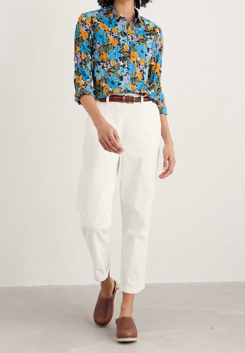 Femme portant une chemise à manches longues à fleurs colorées, rentrée dans un pantalon blanc taille haute avec une ceinture tressée marron et des sabots marron à enfiler.