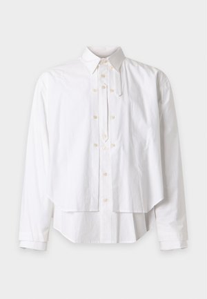 Homme debout portant une chemise blanche à plusieurs couches, un pantalon ample gris foncé et des chaussures marron avec des lacets rouges sur un fond blanc uni.