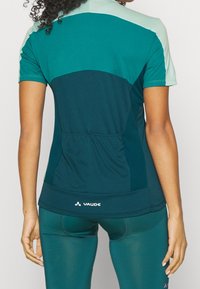 Teal idrottströja med ett nätpanel på baksidan, korta ärmar, rund halsringning och varumärkeslogotypen "VAUDE" vid fållen.