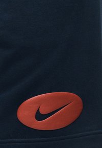 Κόκκινο οβάλ έμβλημα με μαύρο λογότυπο Nike Swoosh σε σκούρο μπλε ύφασμα, που δείχνει μέρος της επιφάνειας και της ραφής ενός ενδύματος.