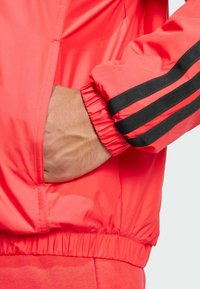 Red windbreaker with black striped accents, elastic cuffs, and a side pocket. Made from smooth, lightweight material.