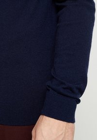 Mörkblå långärmad tröja med synlig ribbad mudd på ärmen och hand mot en enkel ljus bakgrund.