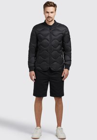 Black padded bomber jacket featuring wave pattern design, zip closure, side pockets, and ribbed cuffs, paired with black shorts and white sneakers.