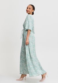 Maxikleid in helltürkis mit Paisleymuster. Kurze Ärmel, Bindegürtel und fließender Stoff. Kombiniert mit pinken hohen Schuhen.