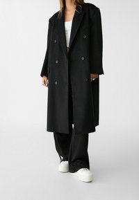 Abrigo negro de doble botonadura con una silueta larga, hecho de una tela suave. Presenta botones grandes y solapas anchas. Combinado con pantalones negros y zapatillas blancas.