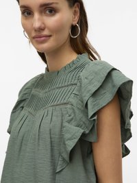 Haut en tissu vert avec des manches volantes, surface texturée et plis verticaux ornés d'un détail de couture décoratif le long de l'encolure.