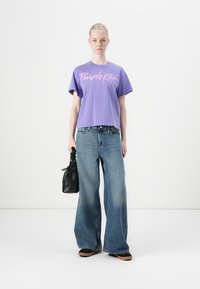 Purple cotton T-shirt with "Purple Rain" text, loose fit, paired with wide-leg blue denim jeans. Black textured handbag in hand.