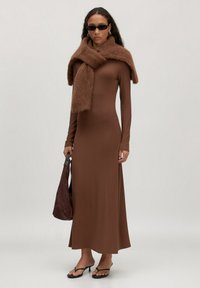 Frau in langem braunen Kleid, mit einem flauschigen braunen Pullover über den Schultern, schwarzen Sonnenbrille, goldenen Ohrhängern, schwarzen Sandalen mit Absatz und einer braunen Tasche.