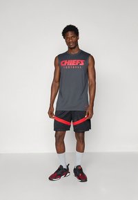 Camisola cinzenta sem mangas com "CHIEFS FOOTBALL" em vermelho, combinada com calções pretos com riscas vermelhas. Usada com meias brancas e sapatos vermelhos e pretos.