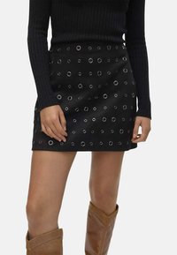 Mini-jupe noire avec oeillets en métal, texture lisse et forme évasée, associée à un haut noir en maille ajusté et des bottes brunes montantes.