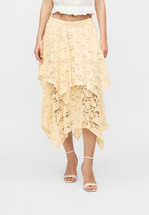 Béžová asymetrická sukňa z čipky, kombinovaná s bielym krátkym topom a béžovými lodičkami s remienkom okolo členka, zobrazená od pásu po chodidlá na jednotnom pozadí.