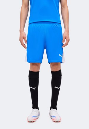 Homme portant un short de sport bleu, des chaussettes noires montantes avec des logos, et des crampons blancs, debout devant un fond uni.