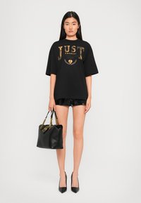Zwarte oversized T-shirt met gouden "JUST CAVALLI" print, gecombineerd met zwarte shorts en een zwarte leren handtas met kettingaccenten.