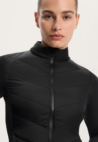 Black zip-up jacket featuring a high collar, padded chevron design on the front, and smooth sides. Made from lightweight, stretchy material.