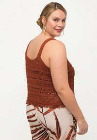 Rostfarbene gehäkelte Tanktop mit breiten Trägern, das ein strukturiertes Muster und einen Fransenrand aufweist. Kombiniert mit leichten, gemusterten Hosen.