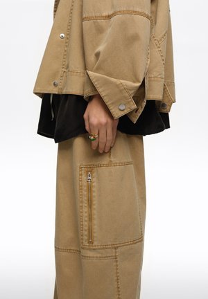 Ręka z kolorowymi pierścieniami spoczywa obok beżowej kurtki i spodni z dużą kieszenią na suwak oraz detalami na zatrzaski na czarnej warstwie materiału.