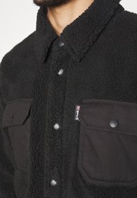 Veste en sherpa noire avec col, boutons-pression sur le devant, deux poches poitrine à rabat boutonné et une petite étiquette de marque en tissu sur la poche gauche.