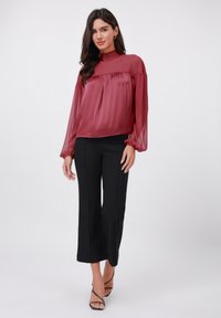 Transparente, burgunderfarbene Bluse mit langen, elastischen Ärmeln und hohem Kragen, kombiniert mit schwarzen, weit geschnittenen Hosen und Riemchensandalen.
