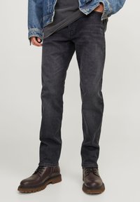 Calças de ganga escuras com um corte slim, combinadas com botas de couro castanhas com atacadores, apresentando uma sola robusta e um acabamento suave.