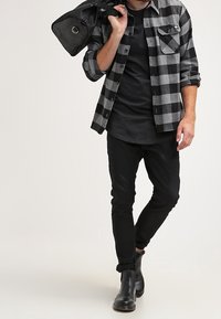 Camisa de franela a cuadros en negro y gris sobre una camiseta negra, jeans skinny negros y botas de tacón negro. Sosteniendo un bolso negro.