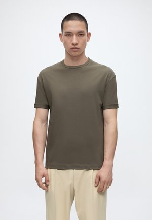 Ung mann med kort hår iført en olivengrønn kortermet t-skjorte og beige bukser, stående foran en ensfarget hvit bakgrunn.