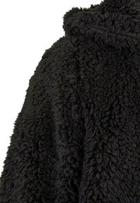 Lähikuva pehmeästä, mustasta, teksturoidusta sherpa-fleece-kankaasta, jossa on pörröinen, kihara pinta, esittäen osan vaatteen olkapäätä ja kaulusta.