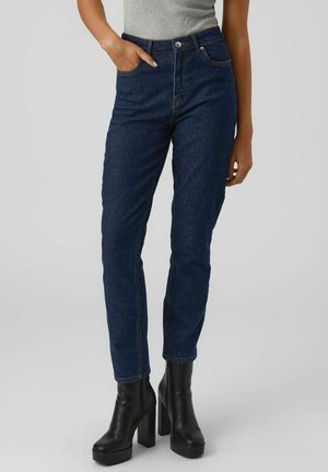 Femme portant un jean taille haute bleu foncé, un top gris sans manches et des bottines noires à talons hauts compensés, debout avec une main dans la poche.