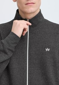 Mørkegrå zip-up jakke med høy krage, med sølvglidelås, ribbestrikkede mansjetter, og en liten hvit logo på venstre bryst.