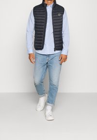 Gilet imbottito nero con colletto alto su una camicia a quadri azzurri, abbinato a jeans azzurri chiari e sneakers bianche.
