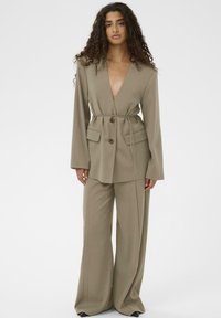 Bēšs pieskaņots komplekts ar platām biksēm un garu, atvērtu žaketi. Raksturīga dziļa V veida kakla izgriezums un jostas saite viduklī. Gluda auduma tekstūra.