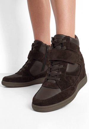 Donkerbruine hoge sneakers met veters, een zijrits en een suède bandje gedragen op blote benen tegen een witte achtergrond.