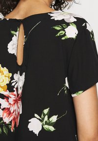Blusa nera con motivo floreale, caratterizzata da maniche corte, scollatura rotonda e un piccolo taglio a forma di chiave sul retro. Tessuto morbido con una consistenza liscia.