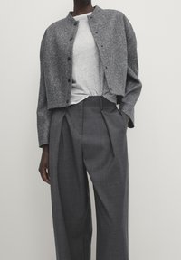 Persoon met een grijze, gestructureerde cropped jas over een lichtgrijs ribgebreid shirt en hoge, geplooide, wijde donkergrijze broek.