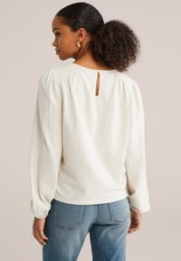 Blusa bianca texturizzata con maniche a sbuffo, dettaglio a occhiello sulla schiena e vestibilità rilassata, abbinata a jeans blu chiaro.