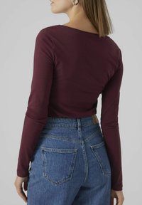 Haut à manches longues bordeaux associé à un jean bleu taille haute avec deux poches arrière et une étiquette en cuir marron sur la ceinture.