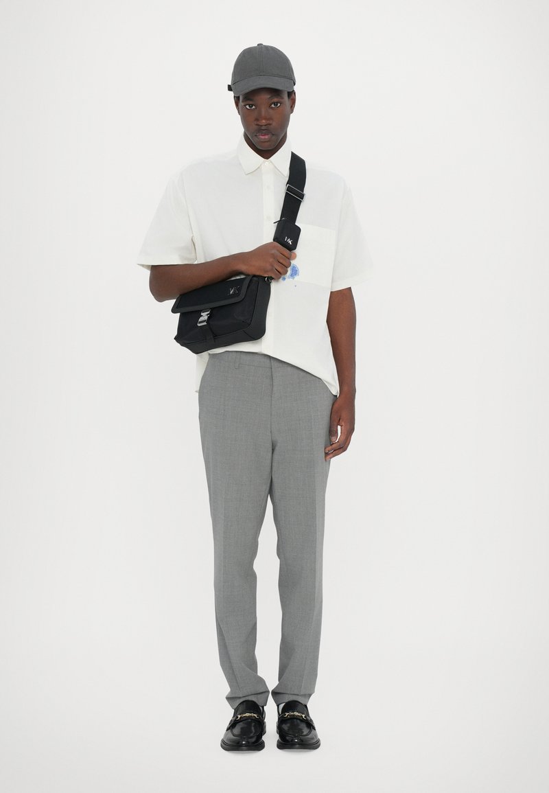 Camicia bianca a maniche corte con taschino; pantaloni grigi a quadri; borsa a tracolla nera; cappellino grigio; mocassini neri con dettaglio a catena.