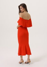 Rochie roșie tip off-the-shoulder cu tiv evazat și volane, siluetă ajustată, textură netedă și fermoar la spate. Pur tată cu sandale cu toc nude.