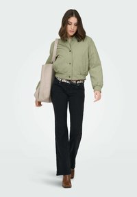 Veste bomber olive courte à texture lisse, avec une fermeture à boutons sur le devant, associée à un jean noir évasé et des bottines marron.