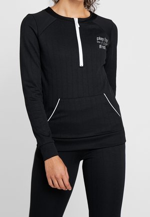 Frau trägt ein schwarzes, langärmliges, geripptes Oberteil mit weißem Reißverschluss und Taschen, auf der Brust ist der Text "play by the rules #not" zu sehen.