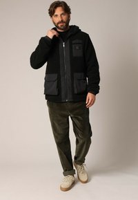 Zwarte fleece-gevoerde jas met een rits aan de voorkant, capuchon en twee voorzakken. Draagt zoals een olijfgroene corduroy broek en beige sneakers.