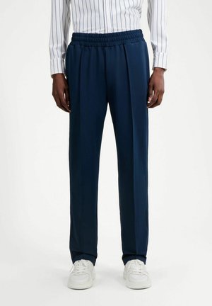 Hombre con pantalones de cintura elástica azul marino, zapatillas blancas y una camisa blanca con finas rayas verticales, de pie contra un fondo blanco.