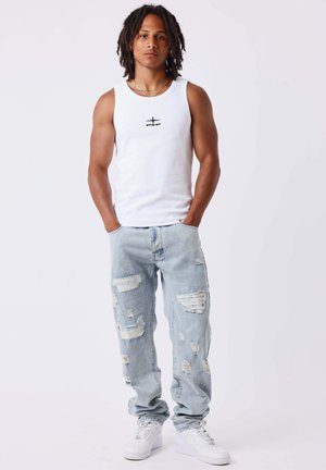 Jeune homme avec des dreadlocks portant un débardeur blanc avec un logo, un jean bleu clair déchiré, des baskets blanches, debout les mains dans les poches.