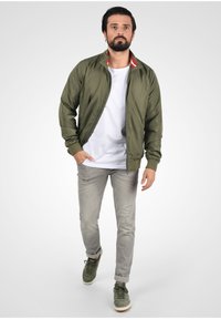 Jachetă bomber verde măsliniu cu fermoar în față, manșete și guler cu nervuri, având accente roșii și albe. Poartă peste un tricou alb și blugi gri.