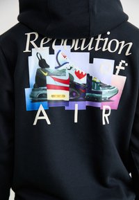 Sort hoodie med et grafisk motiv på ryggen, der viser farverige sneaker-designs og teksten "Revolution of AIR" i beige, med forskellige nuancer i baggrunden.