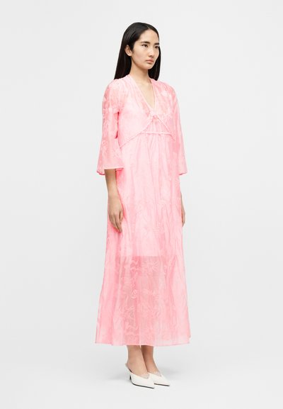 Ροζ μακρύ φόρεμα με floral σχέδιο, μανίκια καμπάνα και ντεκολτέ σε σχήμα V, κατασκευασμένο από διαφανές ύφασμα. Συνδυασμένο με λευκά παπουτσοπέδιλα slip-on.