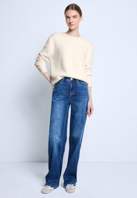 Cremefarbenes, strukturiertes Sweatshirt kombiniert mit hochtaillierten, geraden blauen Jeans. Das Modell steht in lässiger Pose mit leichter Fußbekleidung.
