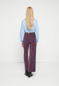 Weit geschnittene lila Cordhosen mit großen Gesäßtaschen, kombiniert mit einem hellblauen Langarmshirt und schwarzen Stiefeln mit Absatz.